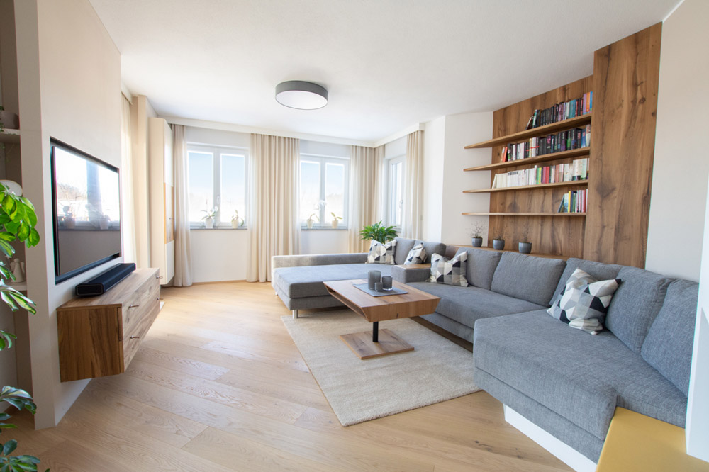 Wohnraum bestehend aus grauem Sofa dahinter ein Holzregal, beigen Vorhängen und Fernseher
